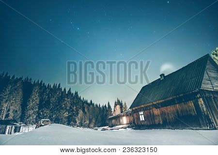 Rural wooden cottage in the mountains with windows glowing in the dark at winter night. Wooden hut on hill moutains.