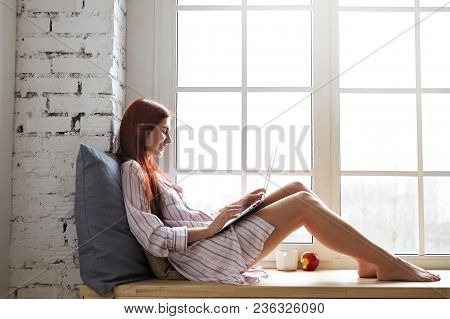 Sideways Shot Of Cheerful Teenage Girl In Striped Shirt Relaxing By Window, Speaking To Friend Via O