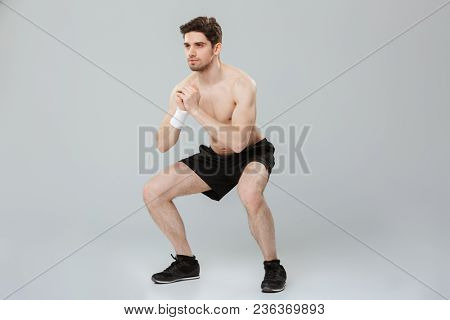 Full length portrait of a confident young half naked sportsman doing squats isolated over gray background