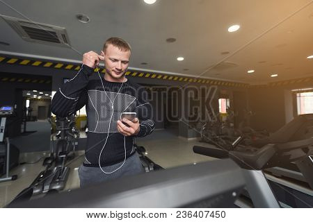 Young Man With Smartphone Running On Treadmill In Gym. Fitness Guy Jogging In Fitness Club, Listen M