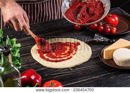 A Male Hand Spreading Tomato Puree On A Pizza Base With Spoon On An Old Wooden Background. Cooking C