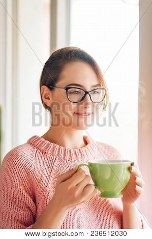 Beautiful Young Girl Standing Next To Window, Holding Cup Of Hot Teaa, Wearing Glasses And Cozy Pull