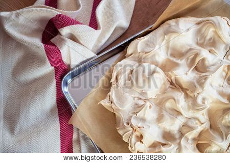 Cooking the meringue base for a Pavlova dessert. Fresh meringue, with crisp golden top, on baking parchment and a stainless steel baking tray. Overhead view on wooden table with cloth.