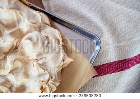 Cooking the meringue base for a Pavlova dessert. Fresh meringue, with crisp golden top, on baking parchment and a stainless steel baking tray. Overhead view on wooden table with cloth.