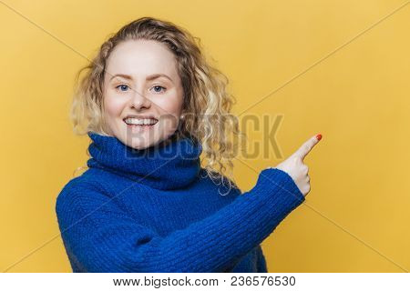 Happy Beautiful Blonde Young Woman Has Pleased Expression, Wears Casual Blue Sweater, Indicates At B