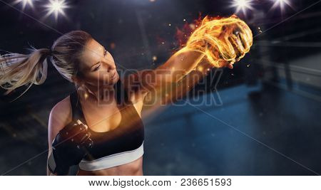 Young blond woman with fire fist hitting the target. Concept of hard work and motivation, boxing arena on background. Very high resolution image
