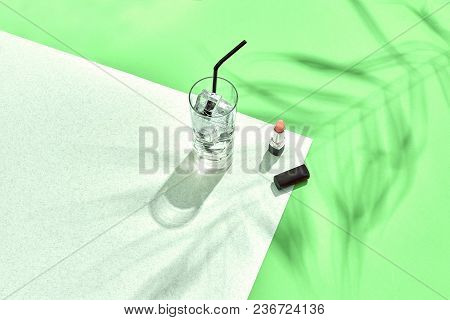 High Angle View Of Lipstick And Drinking Glass With Ice On Table. Green And White Background With Sh