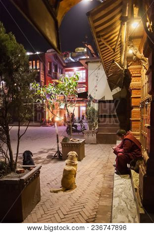 The Buddhist Monk Looks At The Screen Of The Smartphone, Sitting On Stairs Of Houses In An Environme