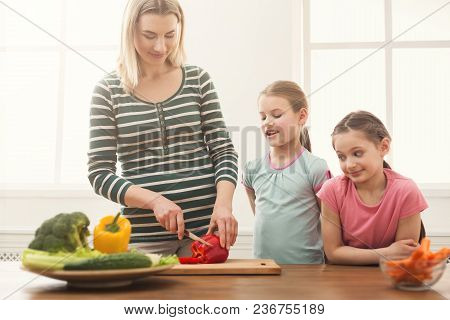 Beautiful Woman With Her Daughters Looks Happy While Cooking Salad In The Kitchen Over Window. Healt