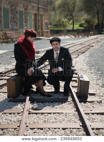 Young Couple With Vintage Suitcase In Deep Thoughts Siting On The Trainlines Waiting For The Next Tr