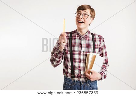 Portrait of an excited young school nerd guy holding book and poinitng pencil up isolated over white background