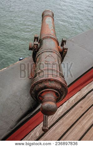 Close-up Of Old Iron Cannon On Wooden Bulwark In Sailboat Docked At Canal Of Amsterdam. Famous For I