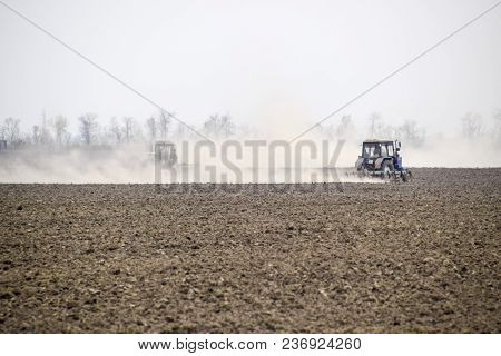 The Tractor Harrows The Soil On The Field And Creates A Cloud Of Dust Behind It.