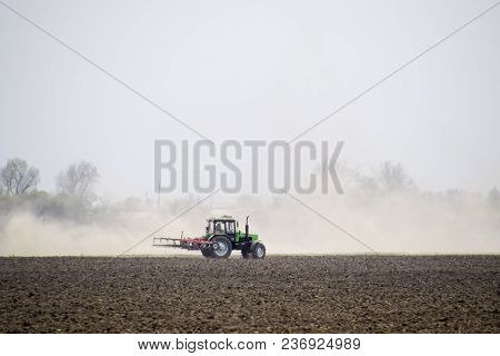 The Tractor Harrows The Soil On The Field And Creates A Cloud Of Dust Behind It.