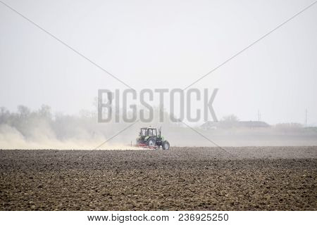 The Tractor Harrows The Soil On The Field And Creates A Cloud Of Dust Behind It.