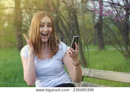 Young Women Girl Happy With Smartphone In Hand. Shy Gets Good News. Blured Green Garden Park.