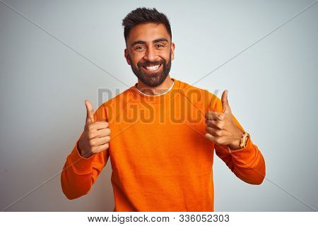 Young indian man wearing orange sweater over isolated white background success sign doing positive gesture with hand, thumbs up smiling and happy. Cheerful expression and winner gesture.