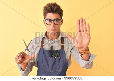 Handsome hairdresser man wearing apron holding scissors over isolated yellow background with open hand doing stop sign with serious and confident expression, defense gesture