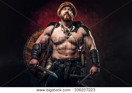 Serious Viking Clad In Light Armor With A Shield Behind His Back Holds A Sword And An Axe. Posing On
