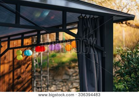 Little Colorful Lampion Lights Hanging On A Pavilion In A Garden On A Sunny Summer Day. Cozy Chairs 