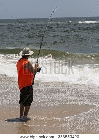 A Man On Fire Island Is Fishing In The Ocean Wearing An Orange Sleeveless Shirt And Standing In On T