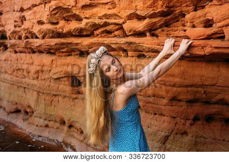 Young Woman With Closed Eyes And Threw Back Head In Crown From Shells Staying Near Red Sand Wall Nea