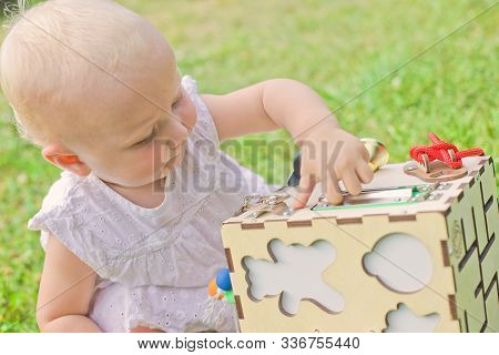Cute Little Girl Is Playing With Busiboard Outdoors On Green Grass. Educational Toy For Toddlers. Gi