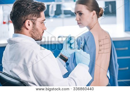 Selective Focus Of Handsome Allergist Holding Syringe Near Patient With Letters On Marked Back