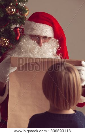 Santa Claus With With List Or Nice Or Naugthy List Paper With Small Boy Near Christmas Tree