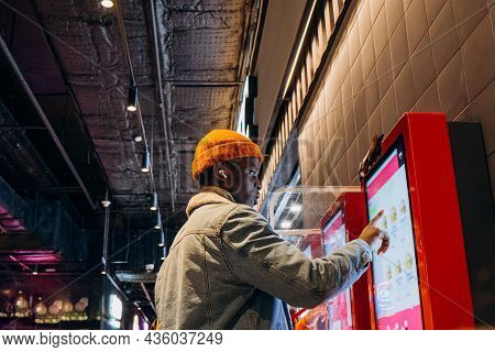 Smiling African-american Man In Warm Denim Jacket With Wireless Earphones Uses Self-service Kiosk To