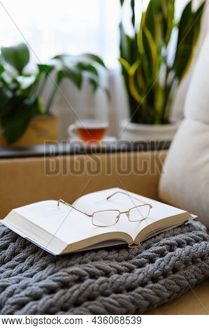 Cozy Reading At Home On A Soft Sofa With A Cup Of Tea. An Open Book With Glasses On A Gray Plaid Aga