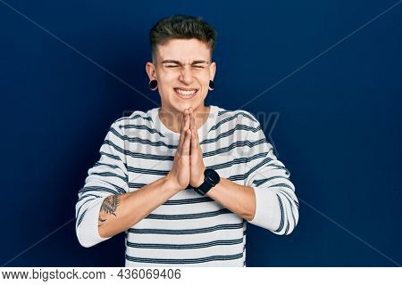 Young caucasian boy with ears dilation wearing casual striped shirt praying with hands together asking for forgiveness smiling confident. 