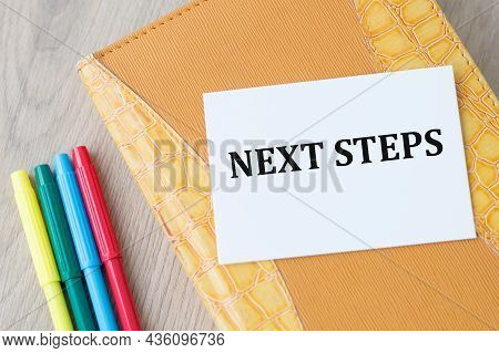 Word Writing Text Next Steps On A White Card That Lies On A Notebook On The Table Next To Colored Ma