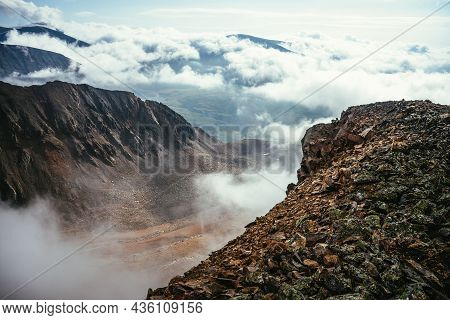 View From Precipice Edge With Sharp Rocks And Snow Cornice Above Clouds To Mountain Valley In Low Cl