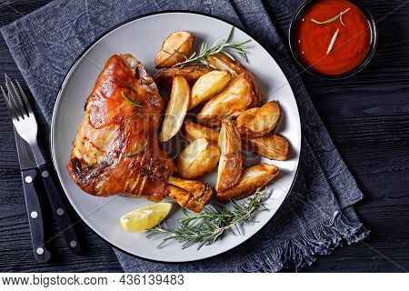 Crispy Roasted Pork Shanks On A Plate With Potato Wedges, Rosemary, Lemon And Tomato Sauce On A Blac