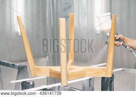 Woman Worker Paints Wooden Chair Base In Transparent Varnish With Spray Gun On Rack Support In Conte
