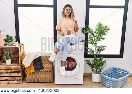 Young hispanic woman drinking coffee waiting for washing machine at laundry room doing happy thumbs up gesture with hand. approving expression looking at the camera showing success. 