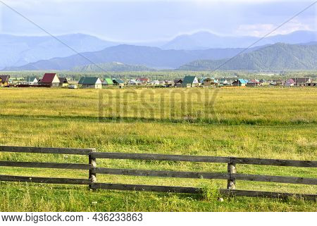 A Fence In A Field, Houses On The Horizon Against The Background Of Blue Mountains In The Evening. A