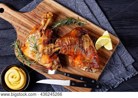 Crispy Roasted Pork Shanks On A Wooden Board With Cutlery, Rosemary, Lemon Wedges And Mustard, Horiz