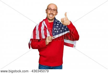 Bald man with beard wrapped around united states flag smiling happy and positive, thumb up doing excellent and approval sign 