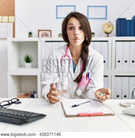 Young doctor woman holding glass of water and prescription pills making fish face with mouth and squinting eyes, crazy and comical. 
