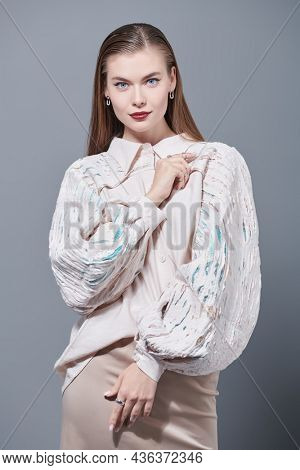 Beautiful girl fashion model with elegant makeup posing in a romantic beige blouse with wide sleeves and a beige straight skirt. High-fashion shoot. Studio portrait on a grey background.