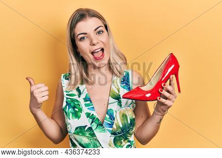 Beautiful caucasian woman holding red high heel shoe pointing thumb up to the side smiling happy with open mouth 