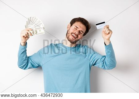 Handsome Man Dancing With Money And Plastic Credit Card, Standing In Casual Clothes Over White Backg