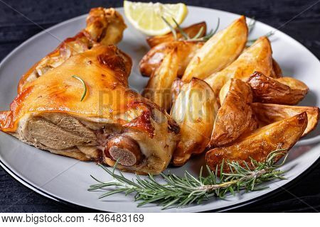 Crispy Baked Pork Shanks On A Plate With Potato Wedges, Rosemary, Lemon On A Black Wooden Table, Hor
