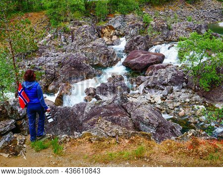 Tourist Woman With Norwegian Flag And Camera Walking Trough Mountains Path In Allmannajuvet Area Sau
