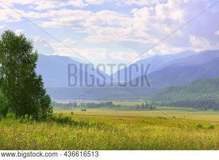 A Green Pasture Against The Background Of Mountains In The Morning Haze Under A Blue Cloudy Sky. Alt