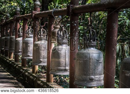 Many Metallic Bells Hanging In A Row On Wooden Pillars Outside In Thai Buddhist Temple. Lined With M