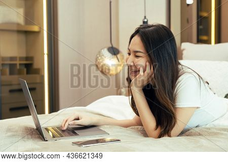 Asian Woman Laying On Her Laptop While Enjoying Herself In Bed In Her Bedroom At Home.