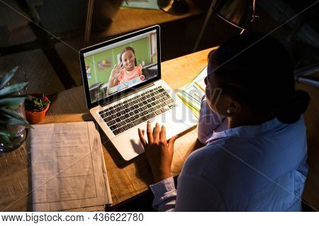 African american woman using laptop for video call, with waving elementary school pupil on screen. communication technology and online education, digital composite image.